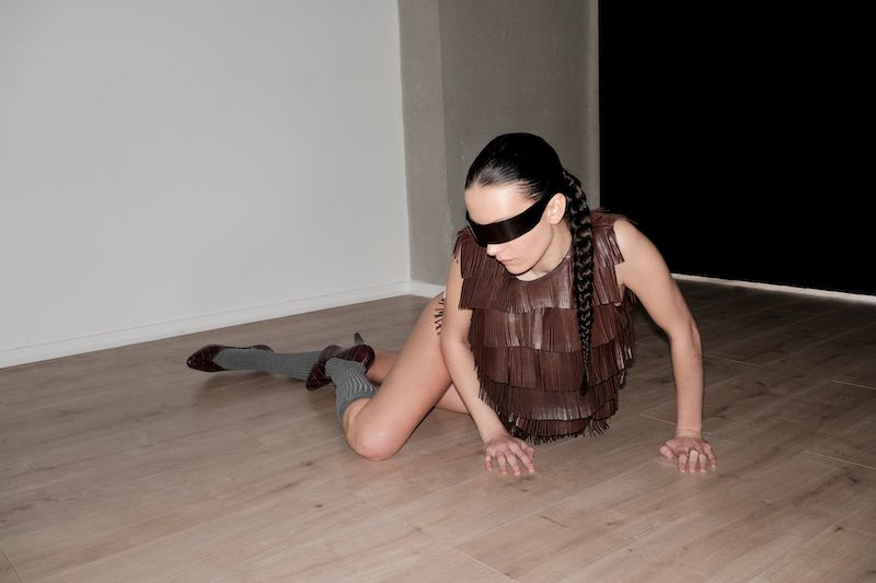 A woman with a braided ponytail and blindfold wearing a fringed top kneels on a wooden floor against a light-colored wall and dark background.