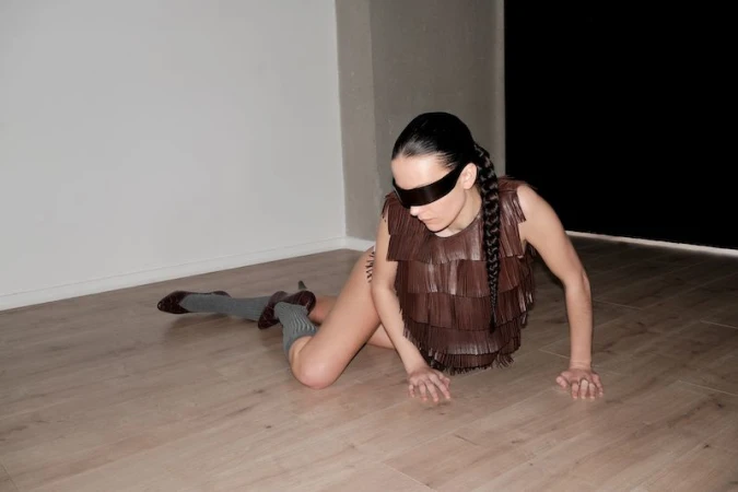 Model wearing a brown fringed top and black eye covering while posing on a wooden floor for Elle Mexico editorial.
