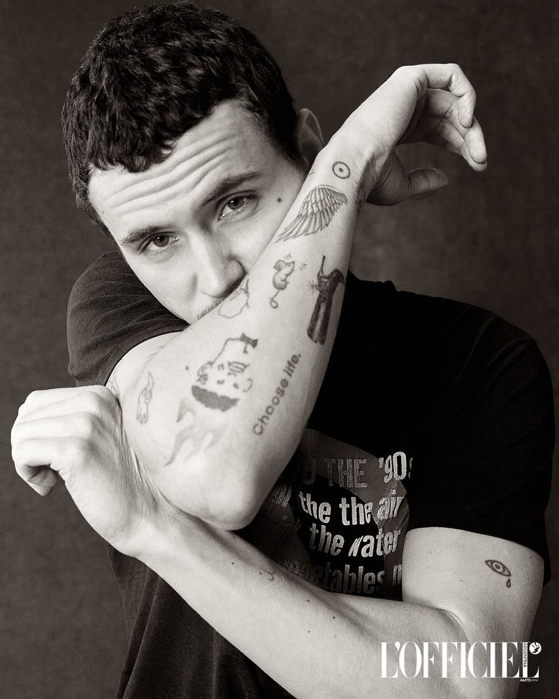 Black and white portrait of a young adult man with tattoos on his arm, dark hair, and a black t-shirt posing indoors.