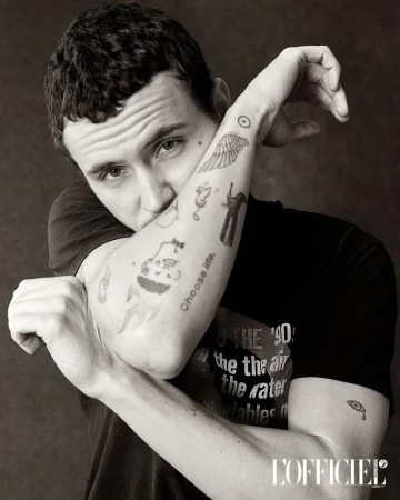 Black and white portrait of a young adult man with tattoos on his arm, dark hair, and a black t-shirt posing indoors.