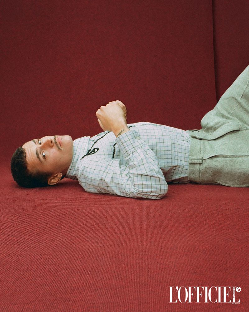 Man in a checkered shirt lies on a red surface, eyes looking up. He wears casual pants. The magazine L'Officiel logo is visible.
