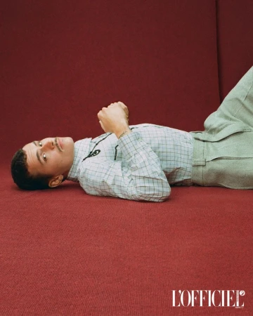 Man in a checkered shirt lies on a red surface, eyes looking up. He wears casual pants. The magazine L'Officiel logo is visible.