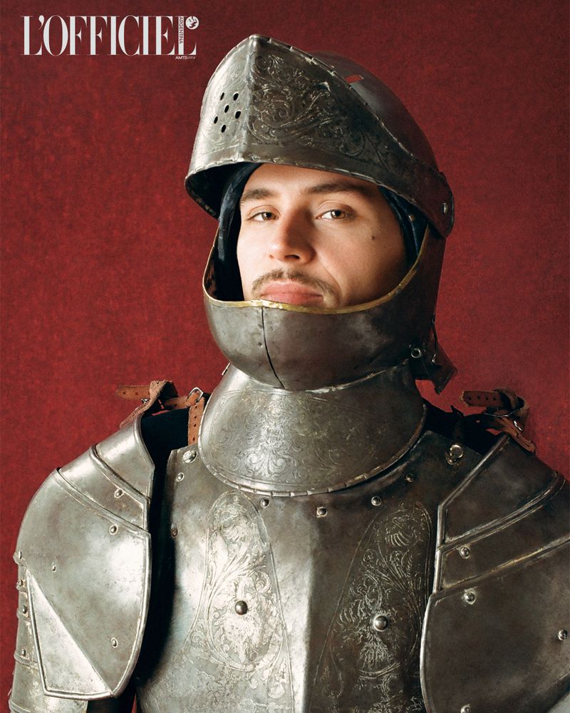 Portrait of a man in vintage armour against a red background. Editorial fashion shot for L'Officiel magazine showcasing historical design.