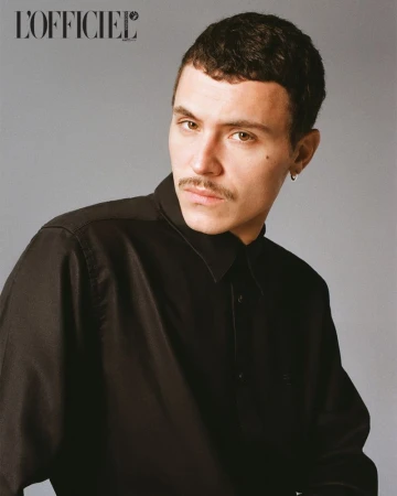 Portrait of a man with dark hair, mustache, wearing a black button-down shirt, set against a neutral grey studio backdrop.