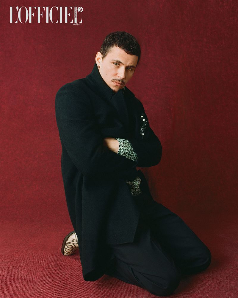 Portrait of a stylish man kneeling with arms crossed, wearing a black coat, patterned shirt, black pants, and patterned shoes against red backdrop.