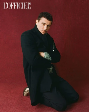 Portrait of a stylish man kneeling with arms crossed, wearing a black coat, patterned shirt, black pants, and patterned shoes against red backdrop.