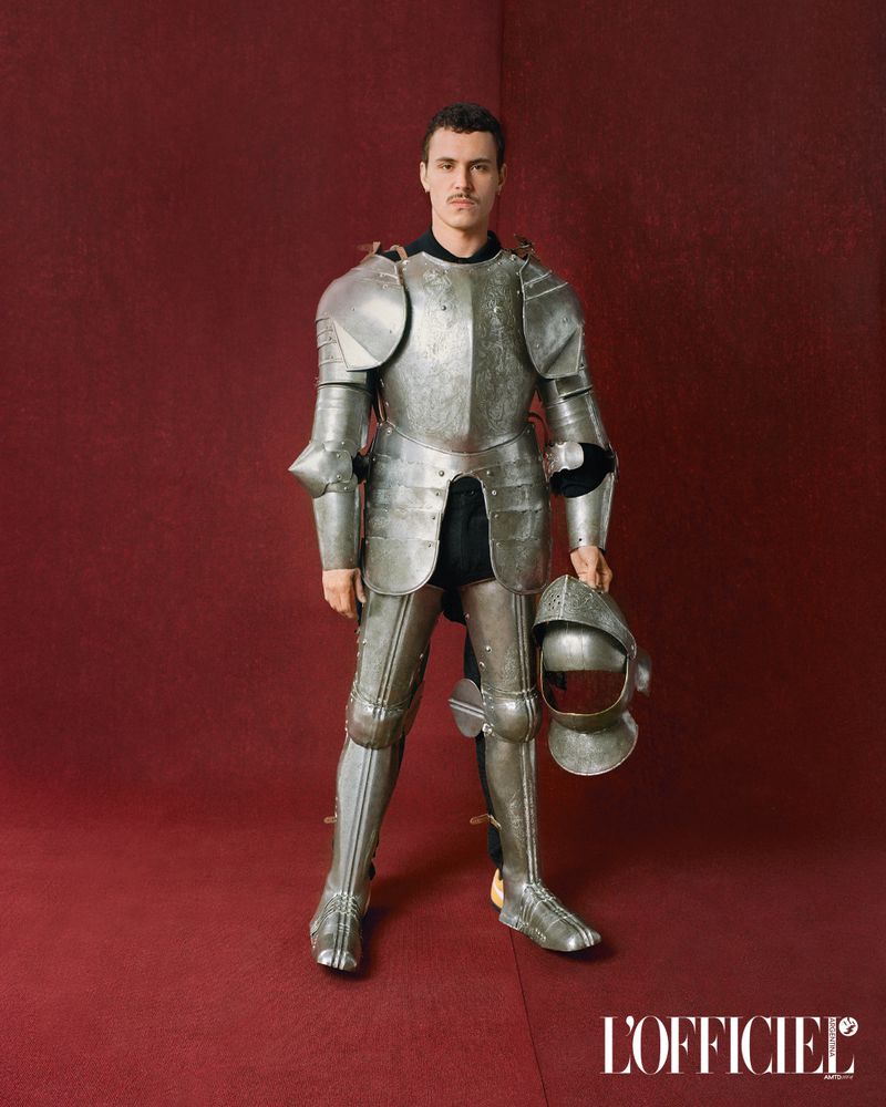 Man in shining knight armor posing indoors against a red backdrop and holding a helmet. Fashion editorial.