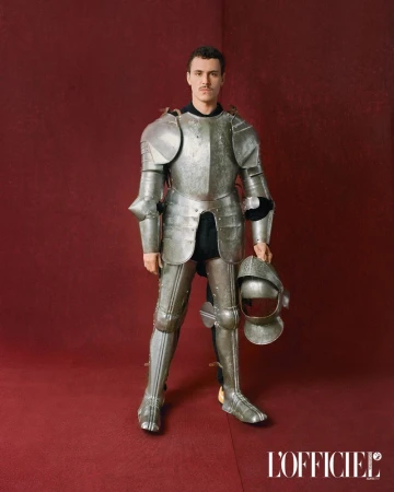 Man in shining knight armor posing indoors against a red backdrop and holding a helmet. Fashion editorial.
