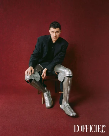 Man poses on chair indoors wearing a blazer and historical armor leggings against a maroon backdrop in a portrait. Editorial.