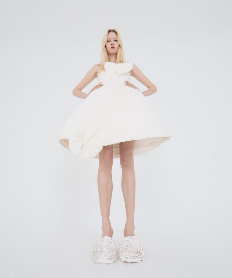 Blonde woman poses in a puffy, white, avant-garde dress and oversized rose-shaped shoes in a bright, high-key lit photo studio setting.