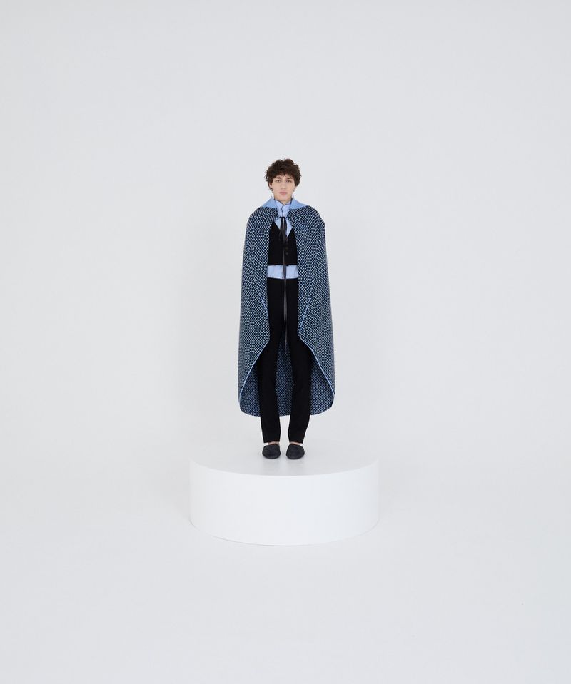 Person stands on platform wearing patterned blue cape and black pants in a brightly lit white studio.
