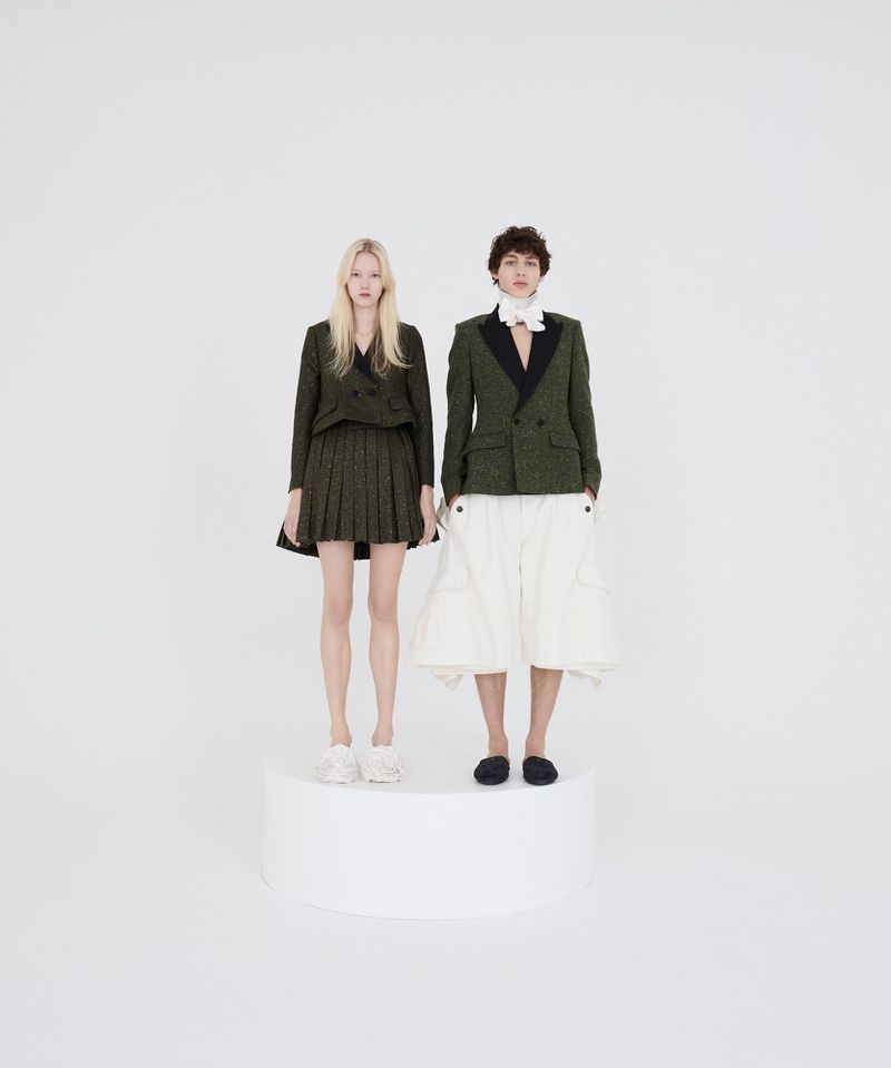 Two fashion models pose on a white platform in matching green tweed blazers and contrasting bottoms in a brightly lit studio.