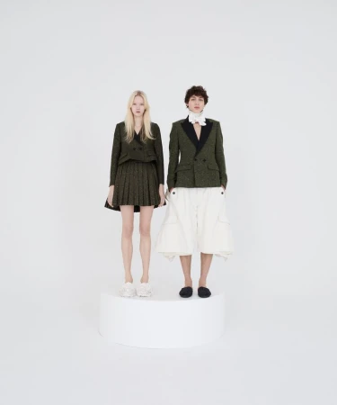 Two fashion models pose on a white platform in matching green tweed blazers and contrasting bottoms in a brightly lit studio.