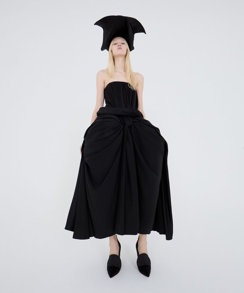 A fair-skinned woman poses in a striking black strapless dress and avant-garde headpiece against a stark white backdrop, showcasing high fashion.