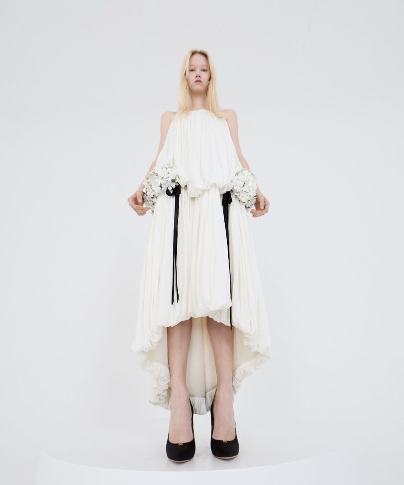 Fashion model in a full-length shot, wearing an off-white pleated dress with floral details and black heels against a white background.