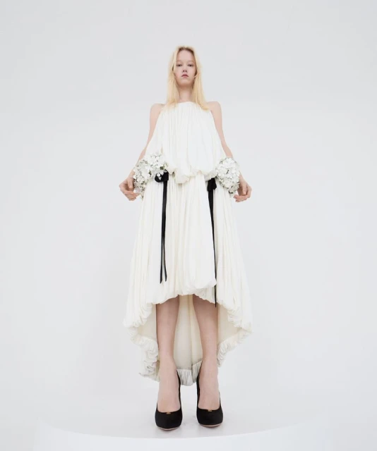 Fashion model in a full-length shot, wearing an off-white pleated dress with floral details and black heels against a white background.