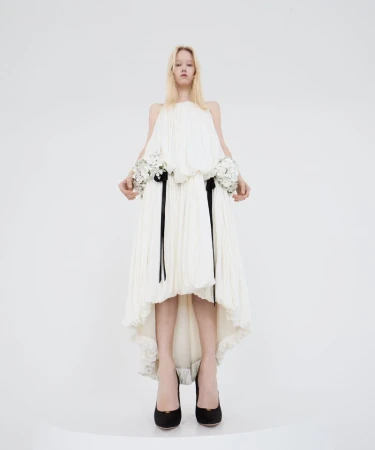 Fashion model in a full-length shot, wearing an off-white pleated dress with floral details and black heels against a white background.