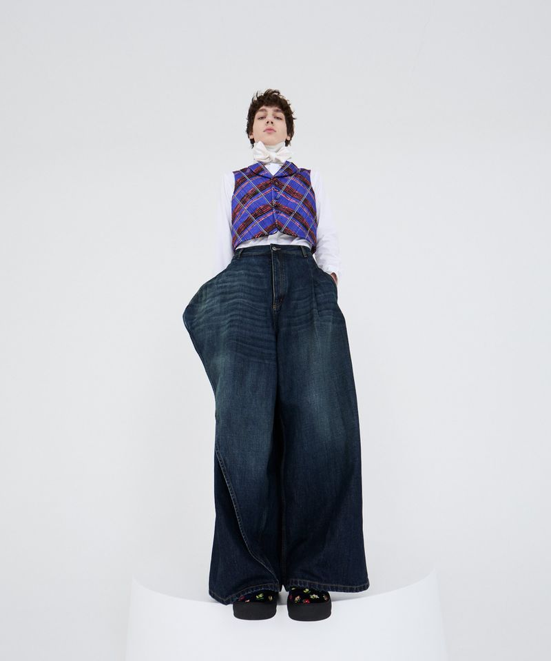 Person in oversized blue jeans and a plaid vest stands on a white platform in a studio setting, showcasing contemporary fashion.