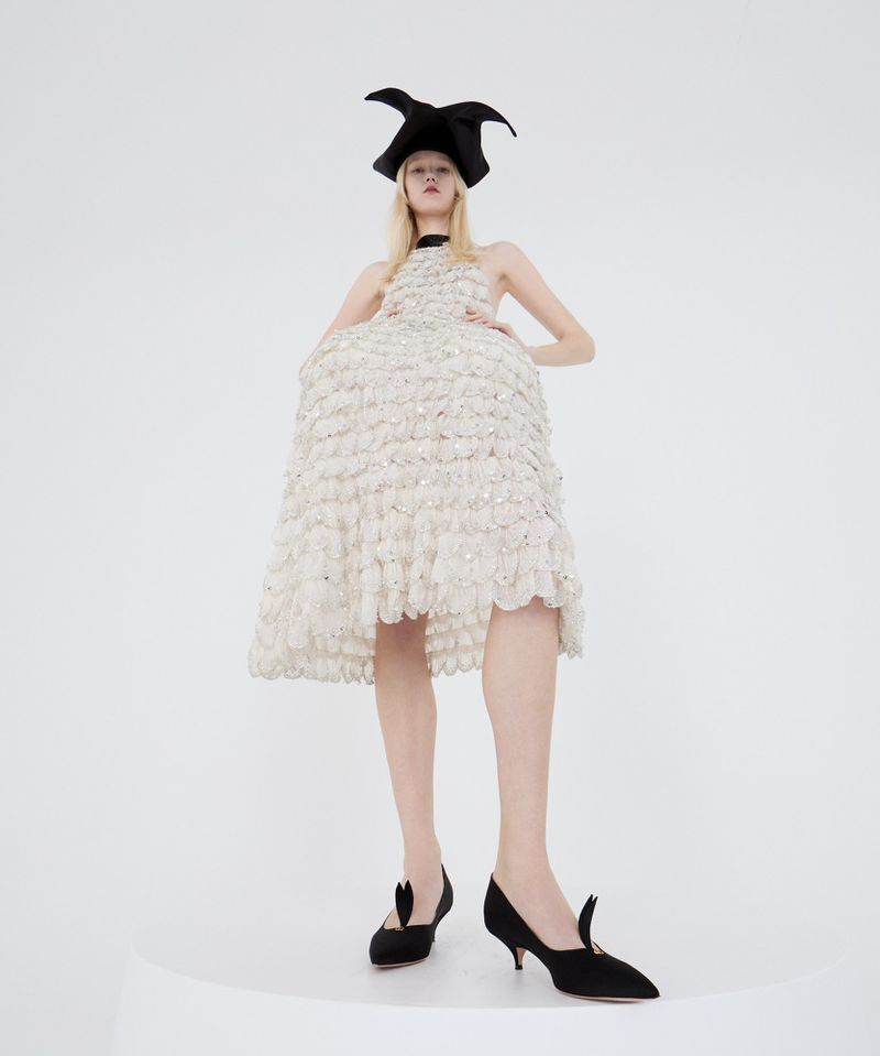 Fashion shot of a slender woman in a white layered dress and black hat, standing on a white surface with black heeled shoes.