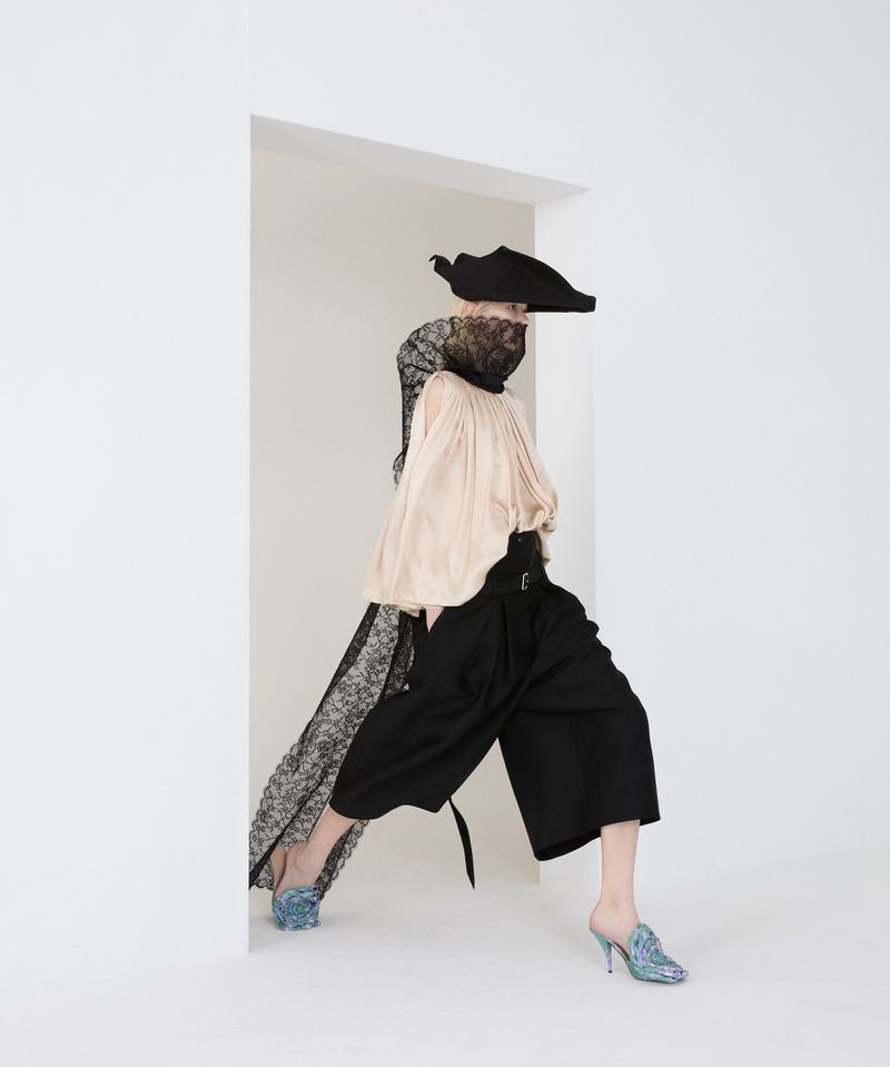 Fashion model in beige top, black culottes, and ornate hat posing in a white doorway. The image highlights studio fashion photography.