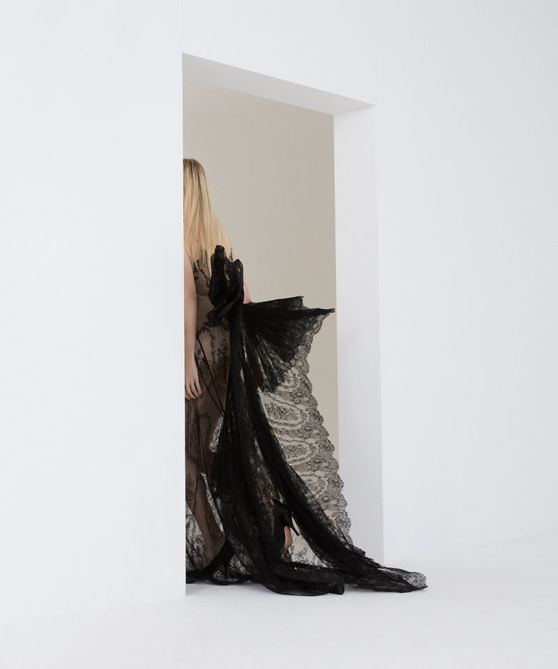 Blonde woman in a black lace gown is seen through a white doorway. Minimalist fashion photography with a clean, bright studio backdrop.