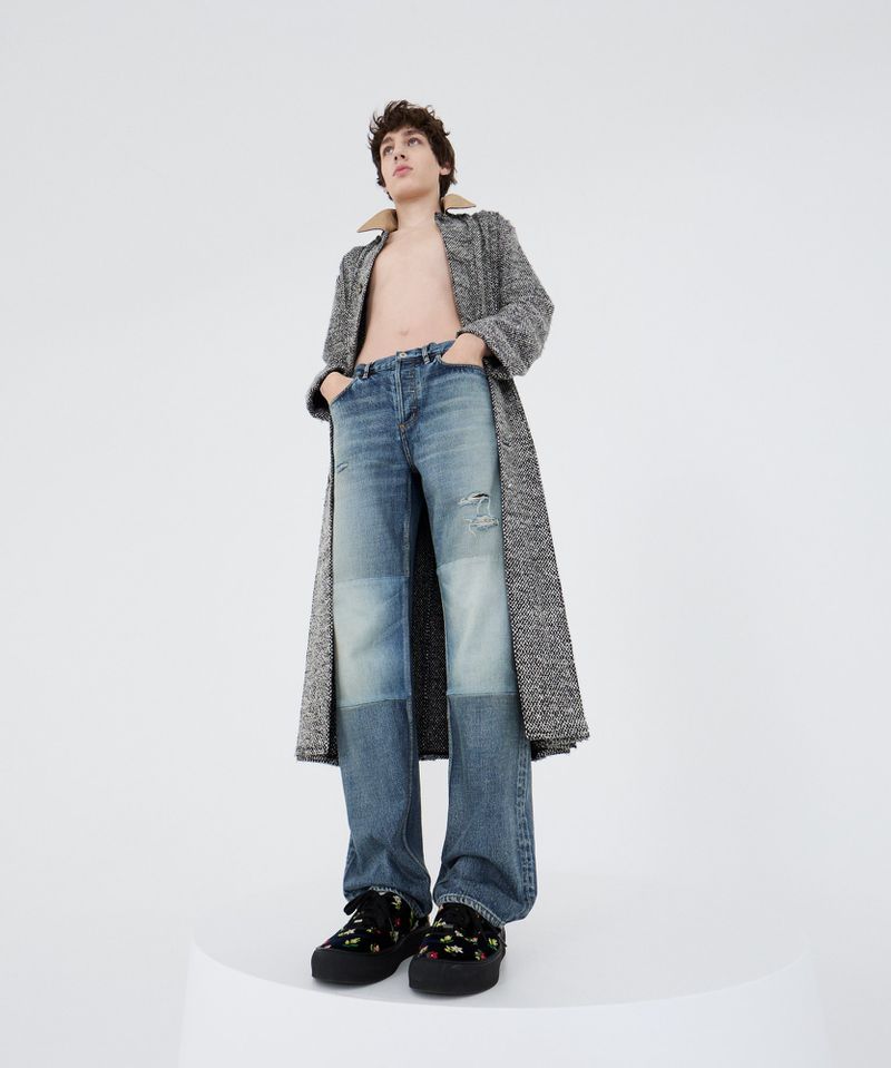 A young adult stands tall in light blue jeans and a gray coat, styled in a light studio with a clean, modern aesthetic.