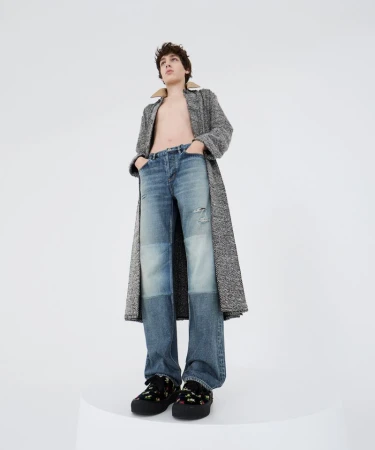 A young adult stands tall in light blue jeans and a gray coat, styled in a light studio with a clean, modern aesthetic.