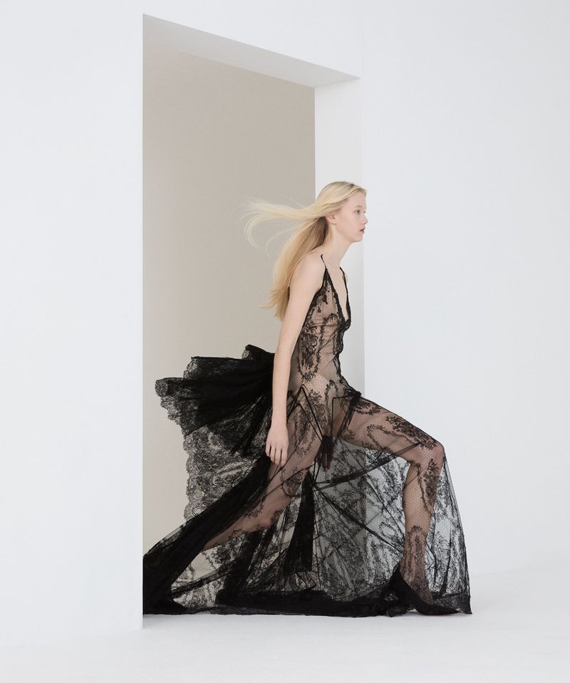 A fair-skinned fashion model with long blonde hair poses in a sheer black lace gown with a train in a minimalist white studio setting.