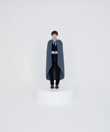 Model wearing a patterned blue cape and dark trousers standing on a white podium against a plain studio background.