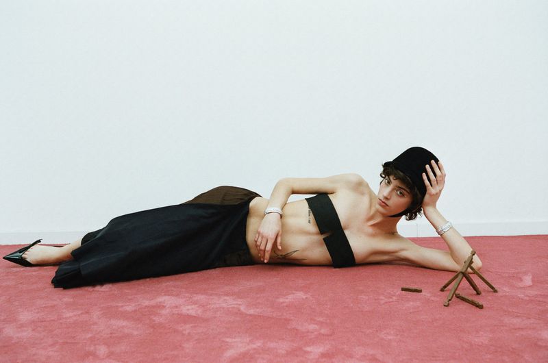 Woman in fashionable black bralette and skirt reclines on a pink carpet next to a small wooden sculpture in a minimalist studio setting.