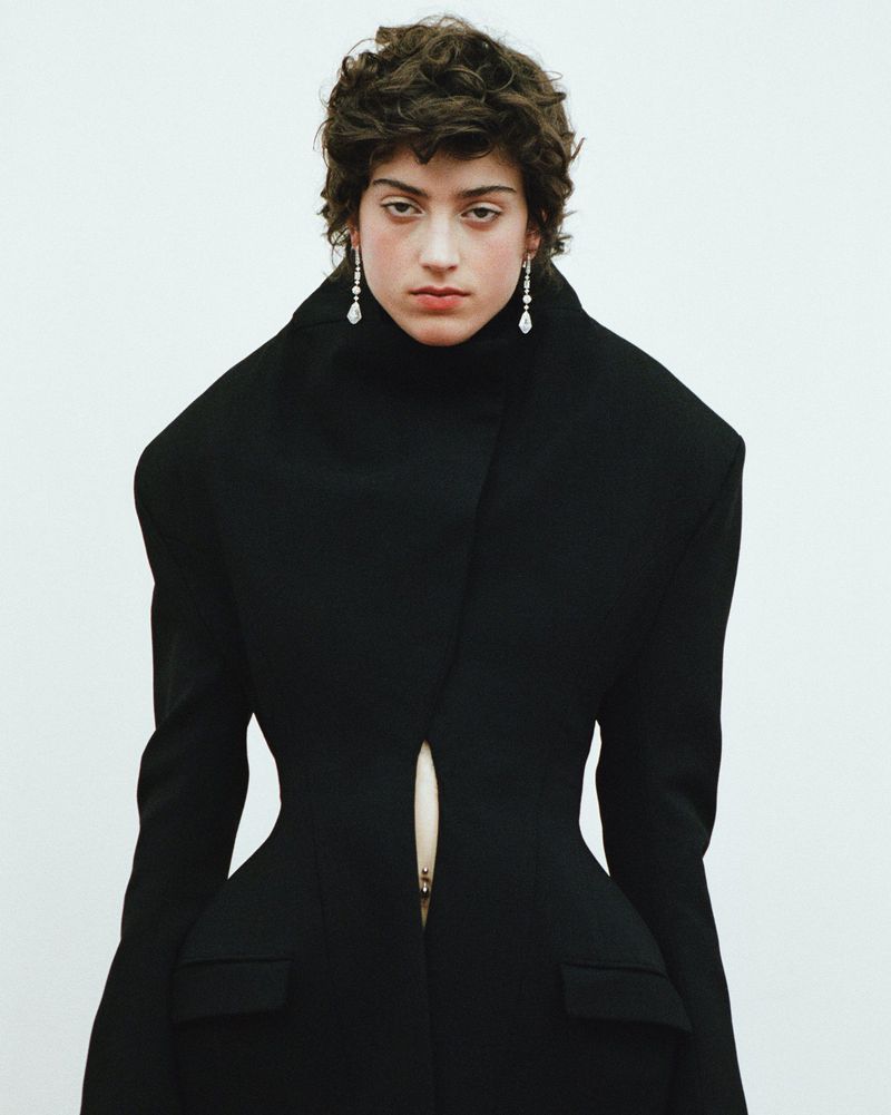 An adult woman with short brown hair poses in front of a white background wearing a long, high-collared, fitted black jacket and drop earrings.