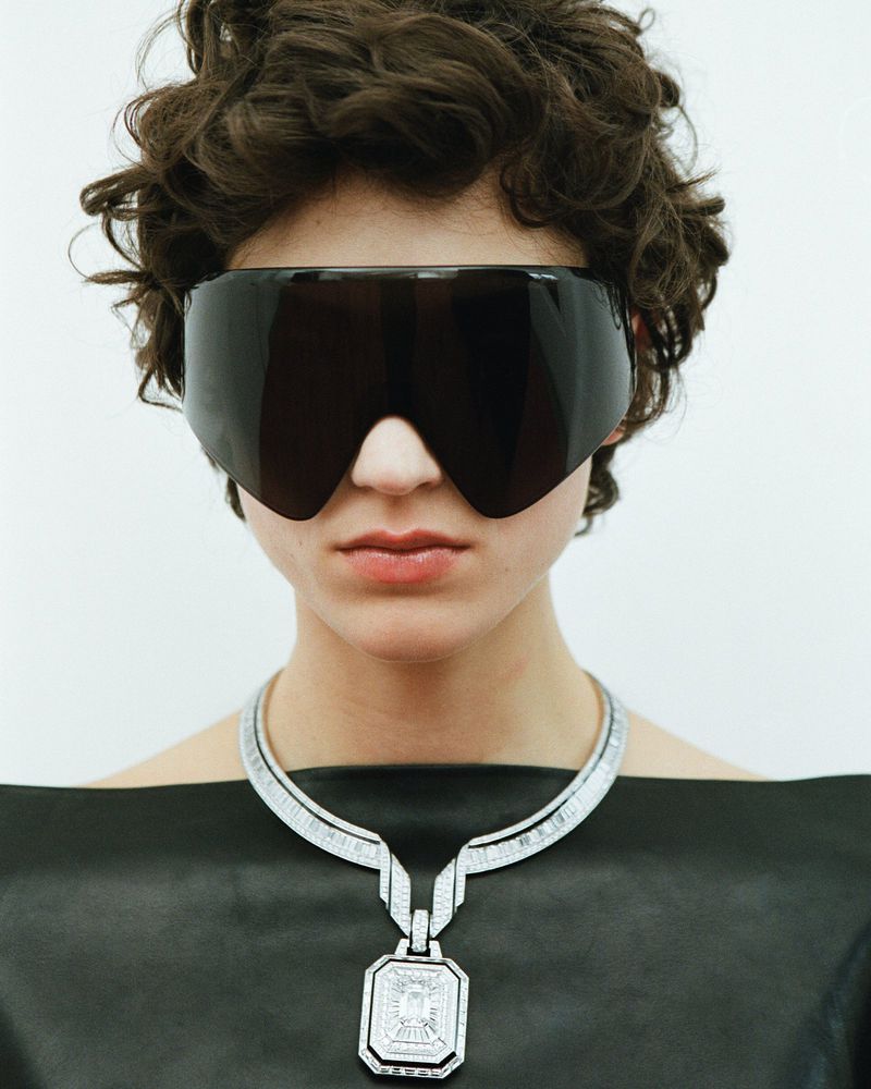 Close-up studio shot of a model wearing sunglasses and a diamond necklace against a white background, showcasing fashion and jewelry.