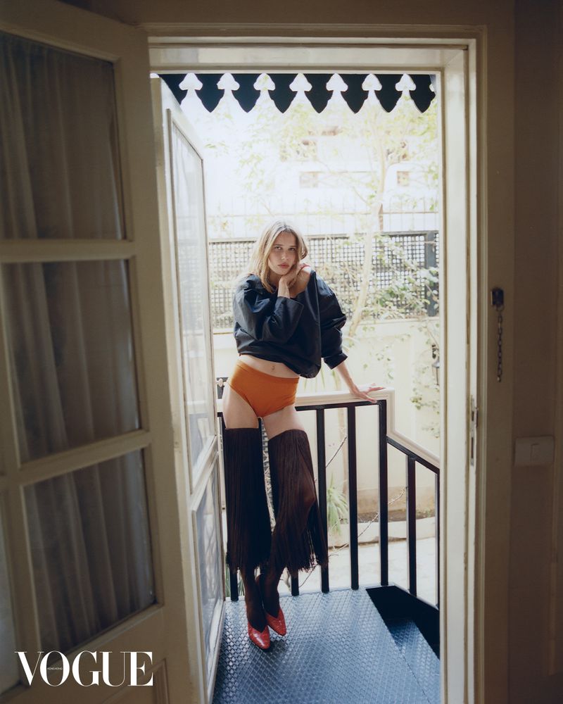 Fashion editorial shot of a woman in orange shorts, black top, and brown fringe boots standing in a doorway with a magazine logo.