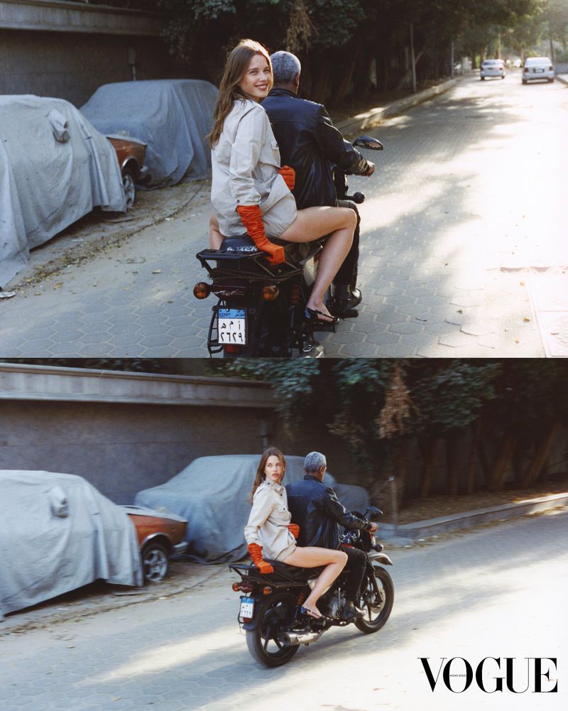 Editorial fashion shot of a young adult female model and adult male riding a motorcycle on a paved street with covered cars in the background.