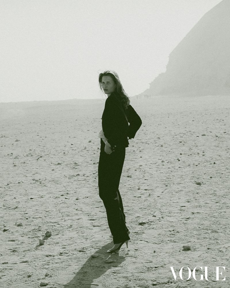 Fashion shot of a woman in a black pantsuit and heels, posing on a tan, rocky plain with a mountain in the background under overcast lighting.
