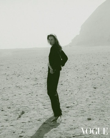 Fashion shot of a woman in a black pantsuit and heels, posing on a tan, rocky plain with a mountain in the background under overcast lighting.