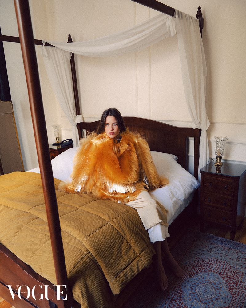 Fashion editorial shot of a young adult female model in a fur coat sitting on a four-poster bed in an indoor bedroom setting.