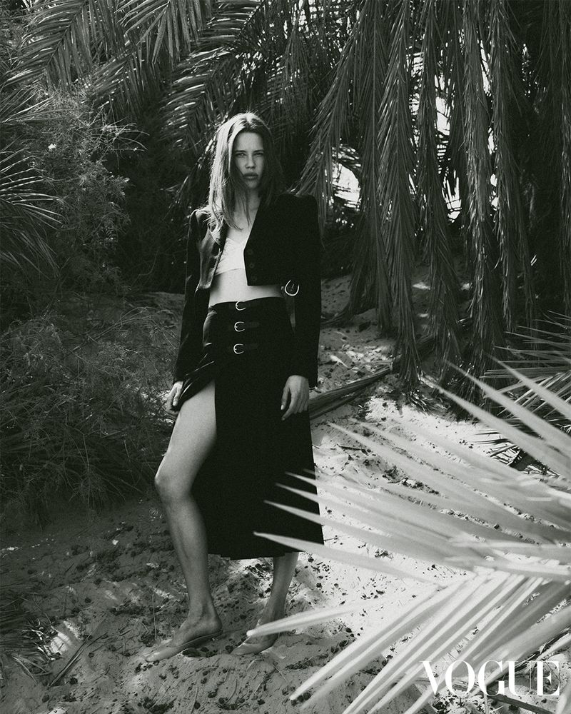 A woman with long brown hair poses in a black skirt and jacket outdoors on a sand dune, framed by palm tree fronds in monochrome.