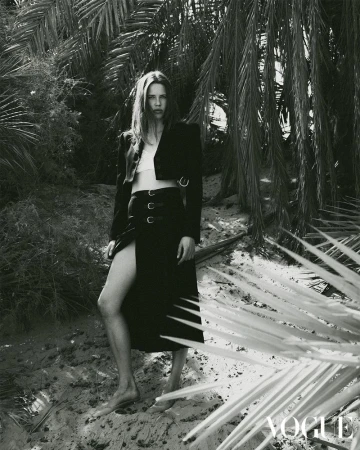 A woman with long brown hair poses in a black skirt and jacket outdoors on a sand dune, framed by palm tree fronds in monochrome.