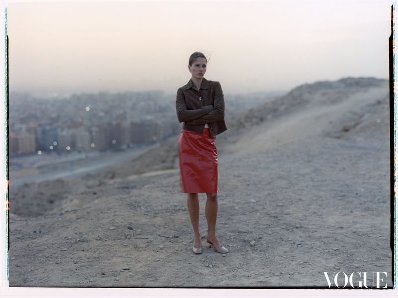 Adult woman stands with arms crossed, wearing red skirt and brown jacket, outdoors on a dirt surface with a city background.