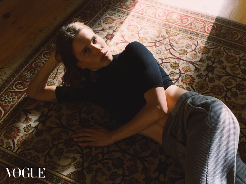 Woman in black top and grey pants lies on a patterned rug in a room with natural light. Fashion editorial shot for Vogue.
