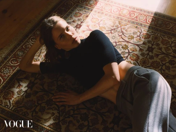 Woman in black top and grey pants lies on a patterned rug in a room with natural light. Fashion editorial shot for Vogue.