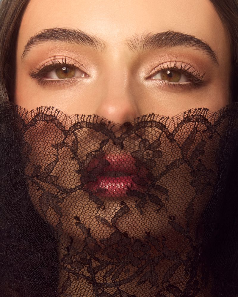 Close-up of a woman's face with brown eyes, wearing dark eye makeup and a dark lace covering her face from the nose down.