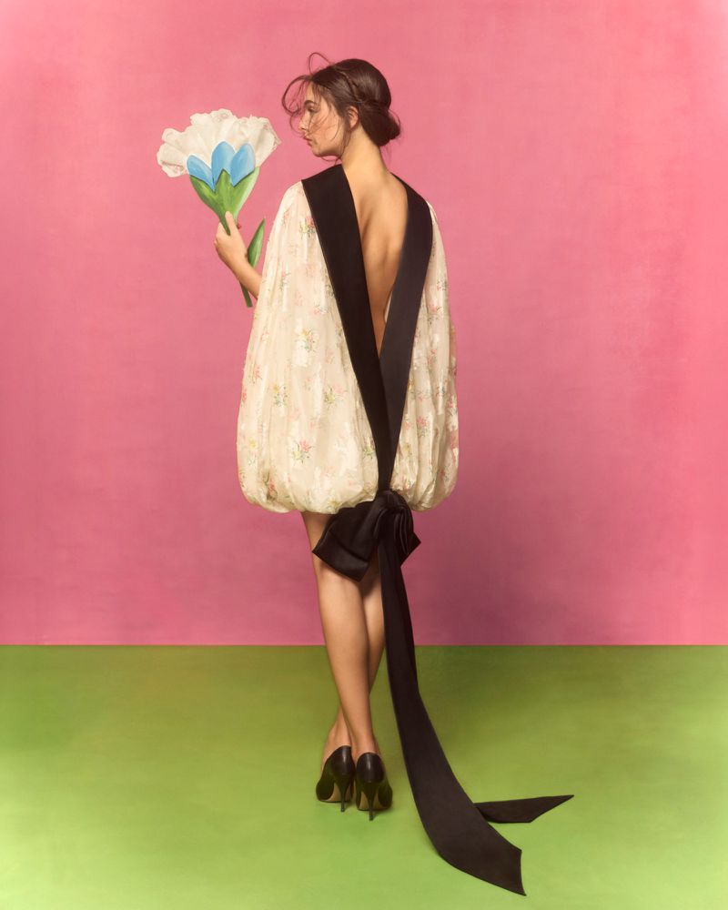Woman in floral dress with black bow poses with large tulip against pink and green backdrop in a fashion style photograph.