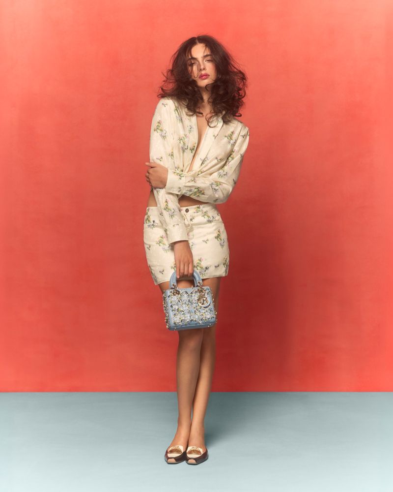 Fashion model in floral blazer and skirt holds purse against coral background; gold shoes complete her polished, stylish look.