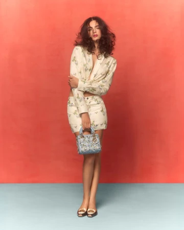 Fashion model in floral blazer and skirt holds purse against coral background; gold shoes complete her polished, stylish look.