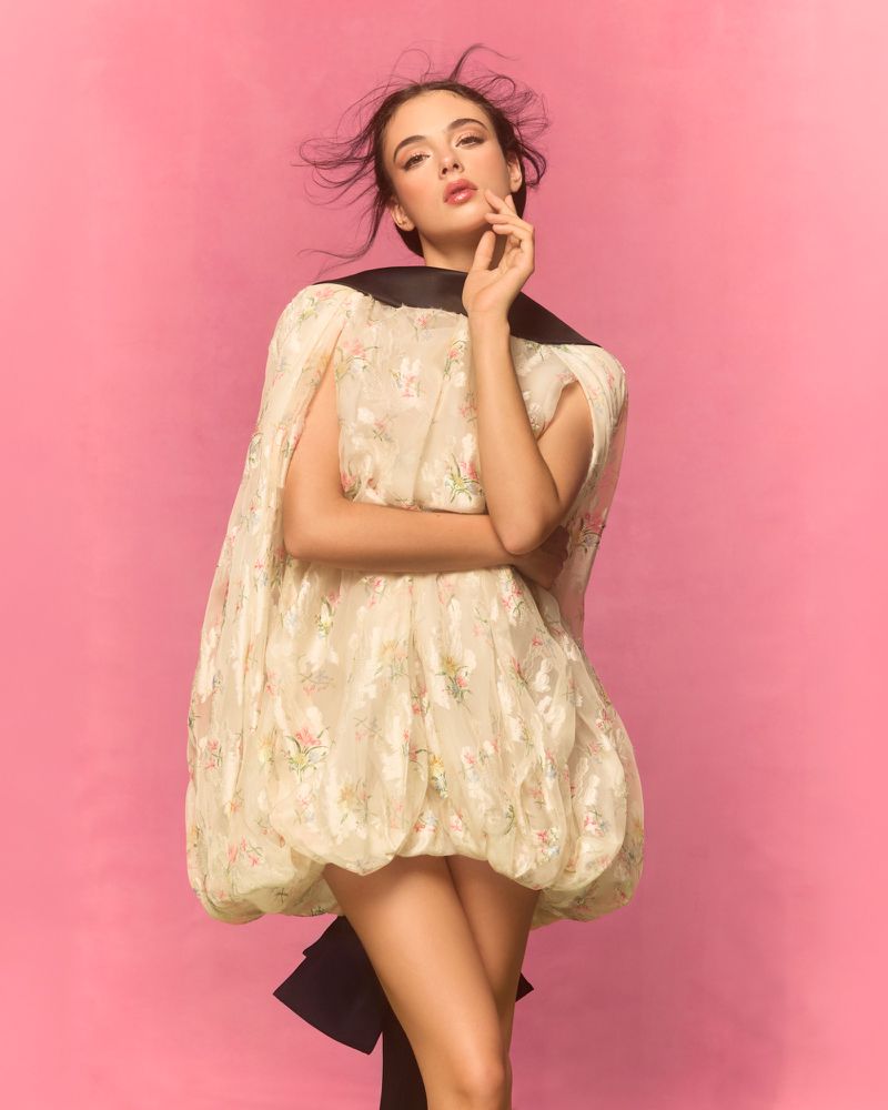 Young woman poses in a cream floral dress with a black accent against a pink backdrop, creating a vibrant fashion portrait in a studio.