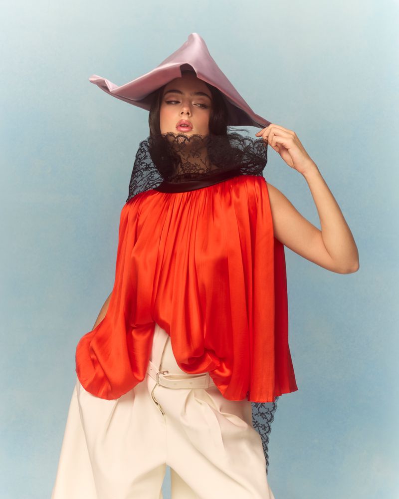 Elegant female model in red top, white pants, and a dramatic lilac hat poses against a soft blue background in a fashion editorial.
