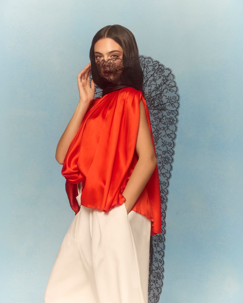 A woman posing in an orange top, white pants, and black lace veil against a light blue backdrop.