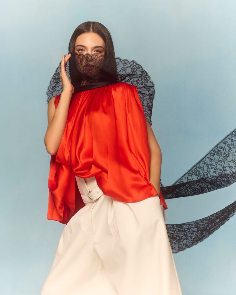 Portrait of a woman wearing a red top, white pants, and black lace veil against a light blue background in a fashion editorial style.